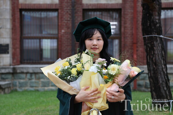 A graduate poses holding a diploma and a flower in front of the College of Humanities.